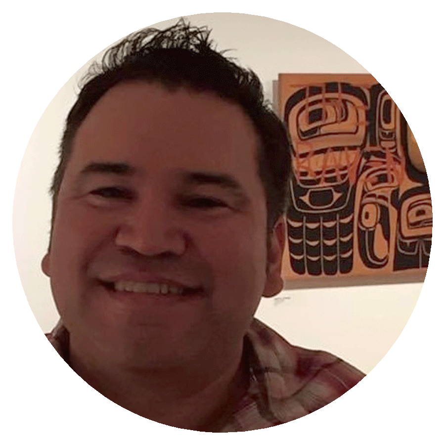 A circularly cropped portrait photograph of a smiling man with short black hair and a plaid collared shirt who is standing in front of a white wall with a piece of wooden, black, brown, and red Indigenous art hanging on it.