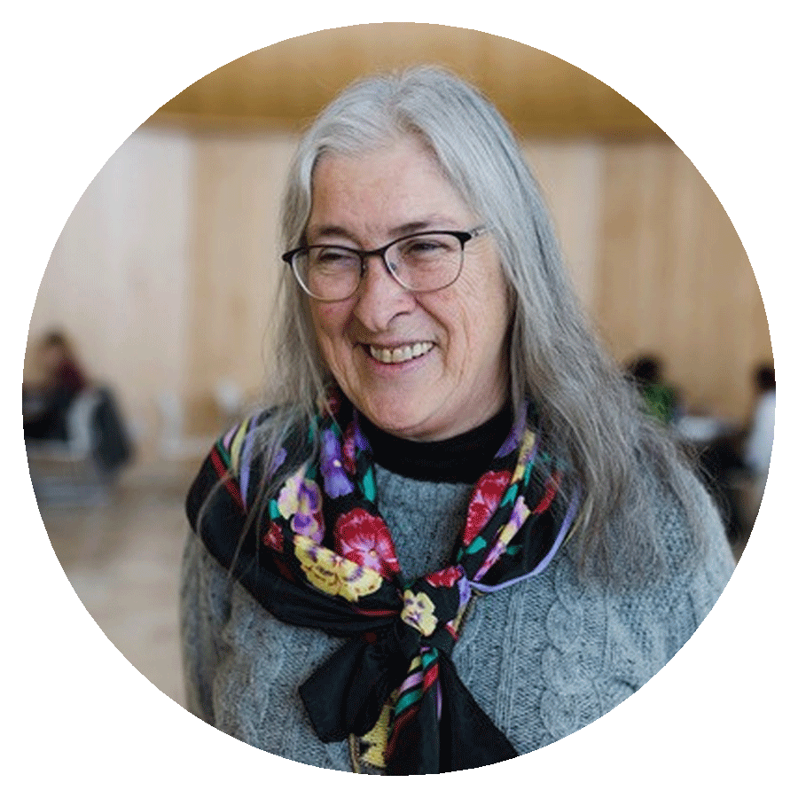 A circularly cropped portrait photograph of a smiling woman with long grey hair, glasses, a colourful scarf, and a grey knit sweater who is standing in a large light brown room with people sitting at tables in the background.