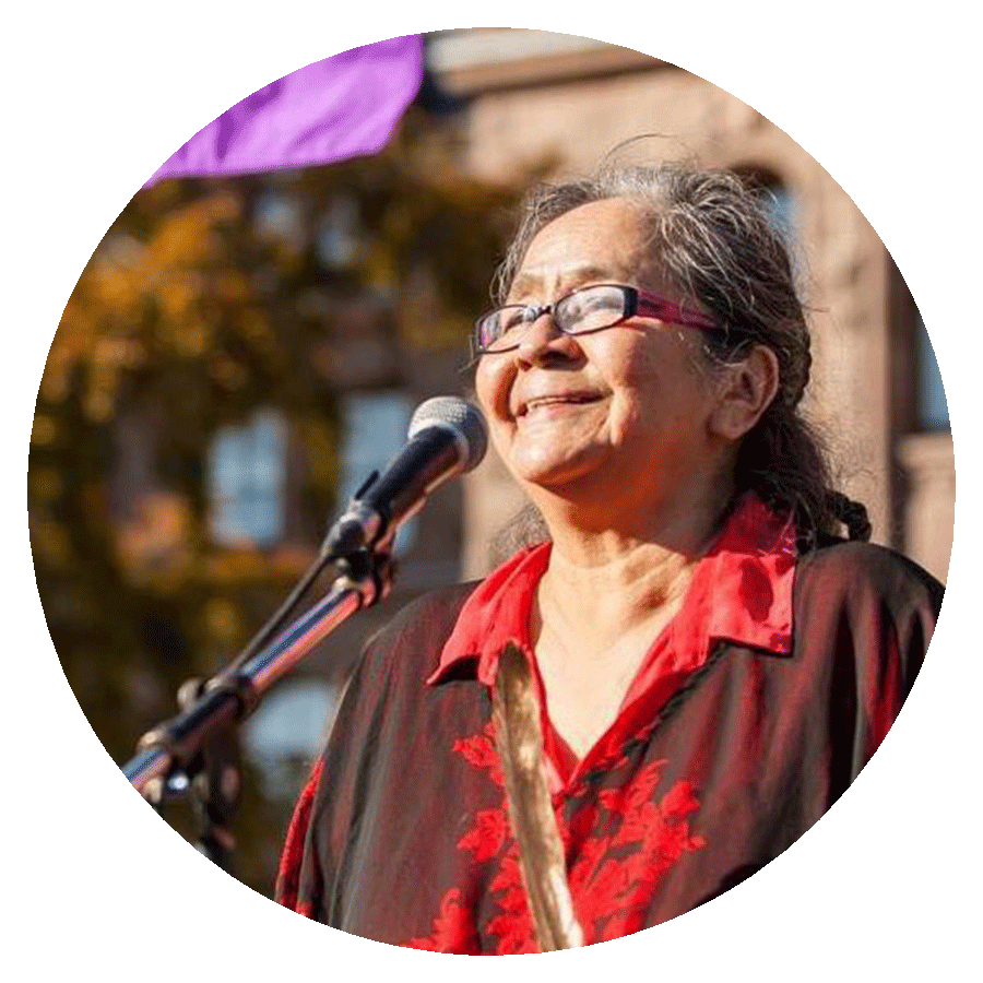 A circularly cropped photograph of a smiling woman with long grey hair, glasses, and a red shirt who is standing outside on a sunny day in front of a microphone.