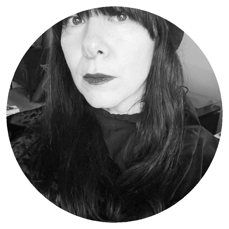 A circularly cropped black and white portrait photo of a woman with black clothing, long dark hair, and dark lipstick who is standing in an interior space.