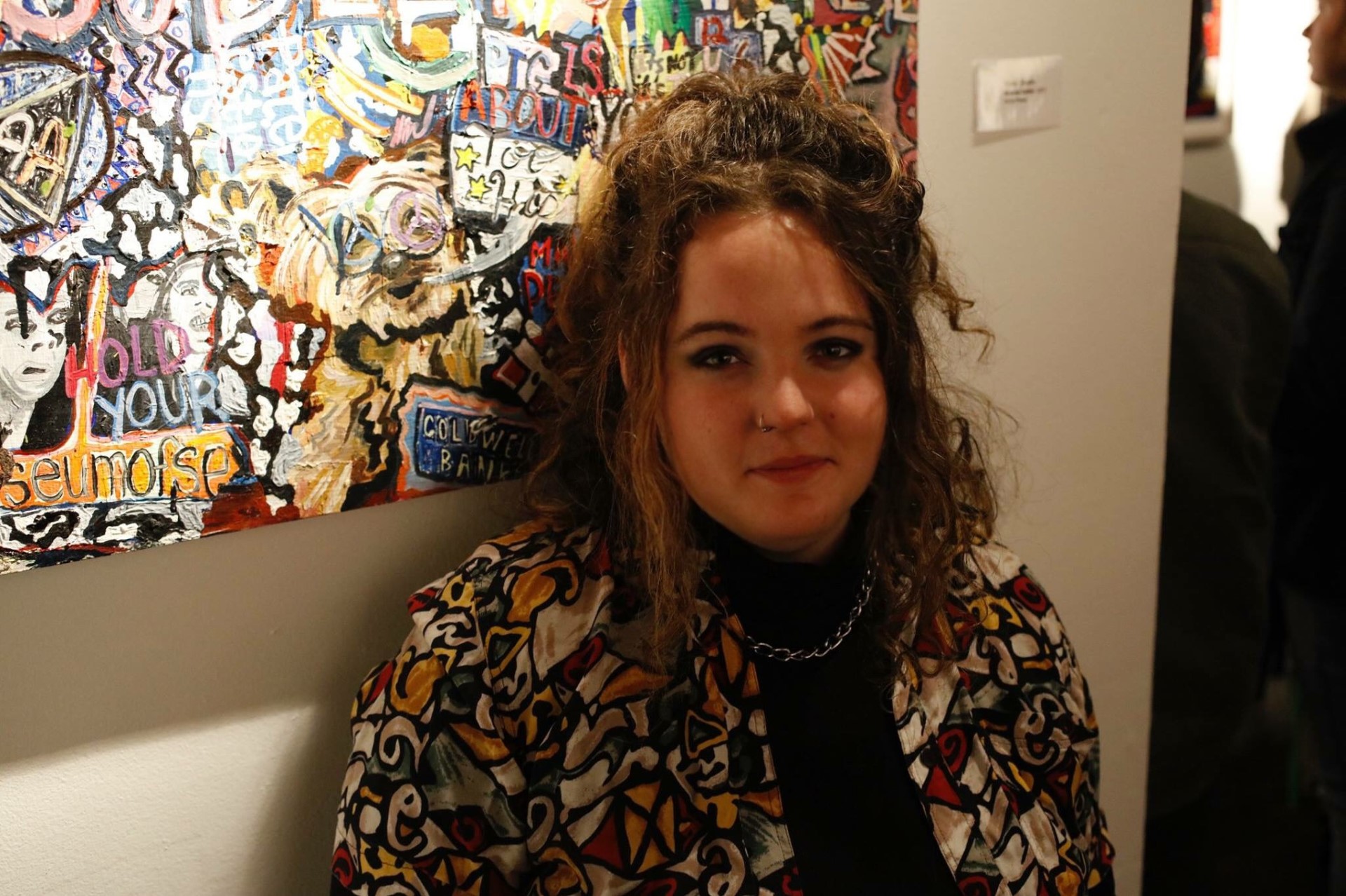 A portrait photograph of a woman with curly light brown hair standing in front of an abstract painting.