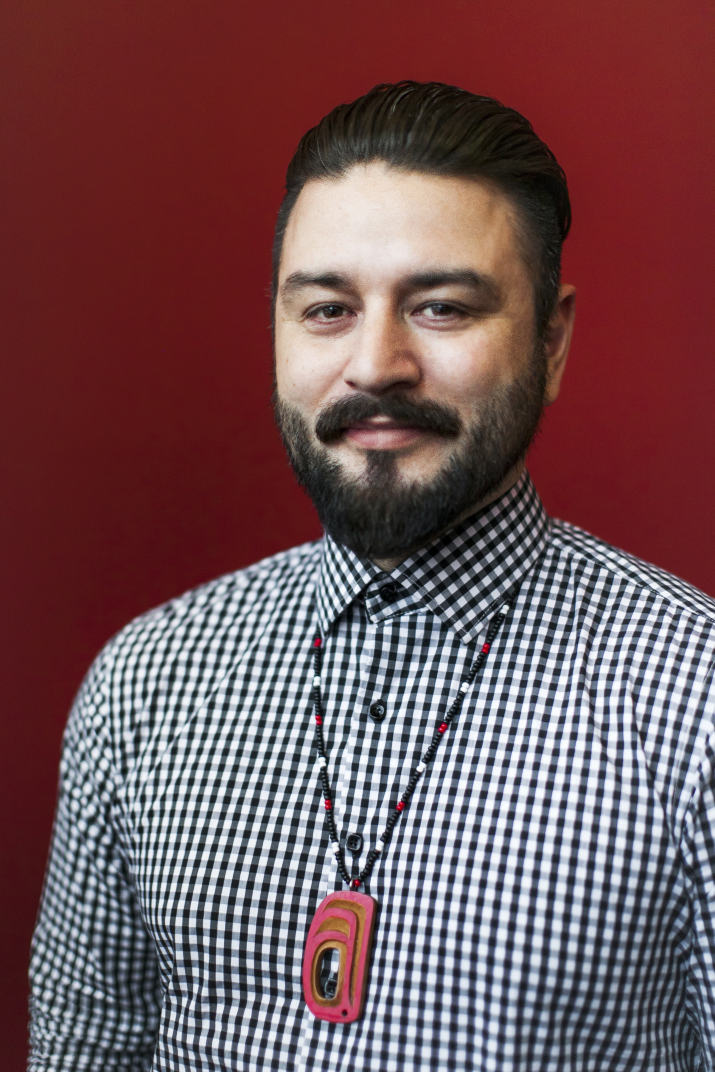Smiling man with short dark hair, moustache and beard with b/w checked shirt and beaded necklace