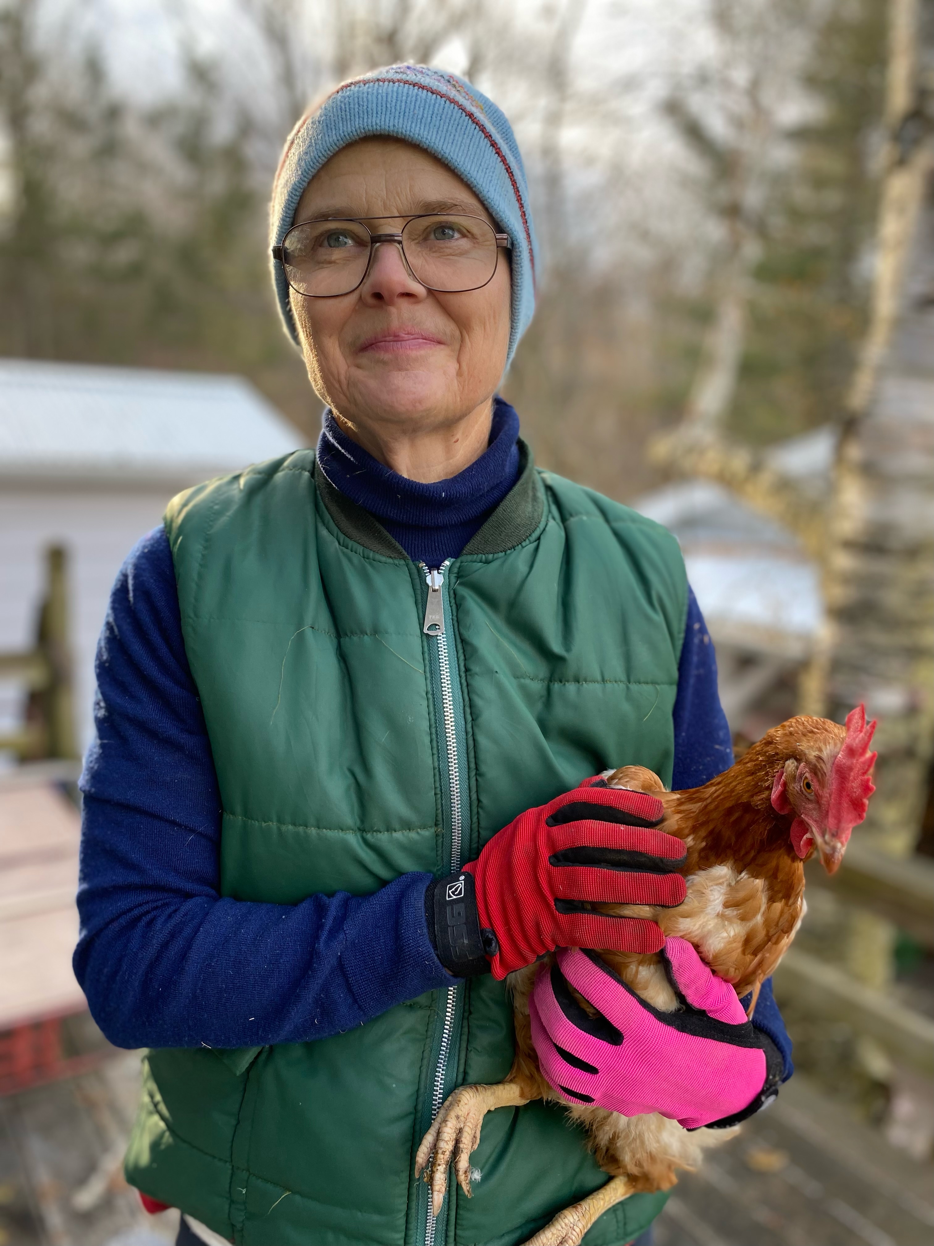 A photograph of a woman with glasses and colourful winter clothing standing outside in snowy rural area holding a chicken.