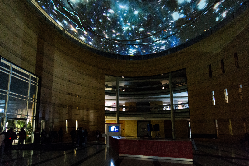 A colour photograph of a university lobby space at night where a large digital video projection is being shown on the building's domed ceiling. The video is of an animation of a starry night sky with line drawings of animals moving around between the stars.