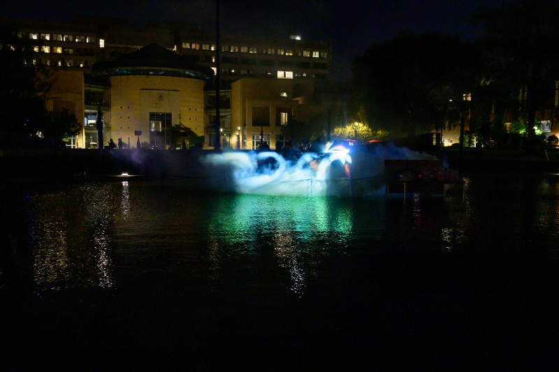 A colour photograph taken at night of a video projected onto fog of a group of white swans. The projection is located above an artificial pond at a university campus.