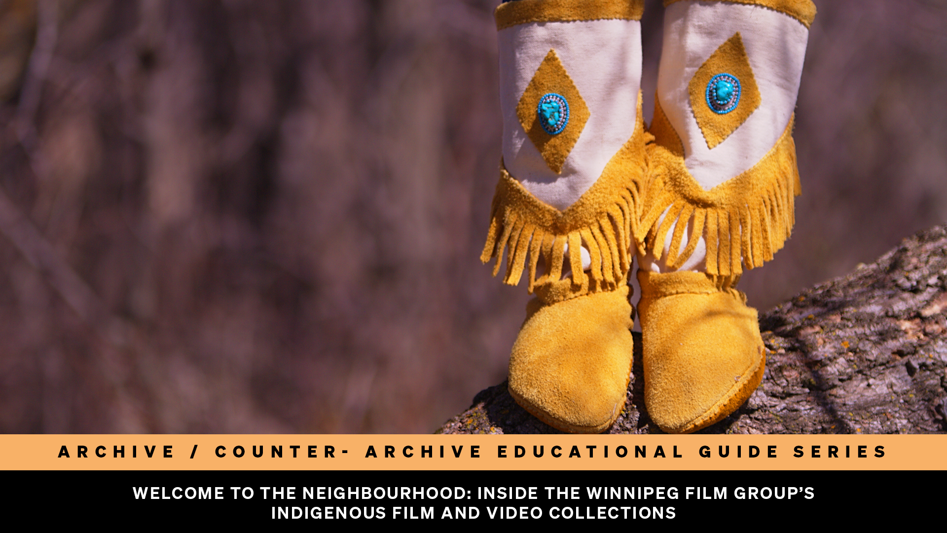 A close-up of a person’s feet wearing yellow moccasins with fringe and decorative blue diamond-shaped designs. They are standing on a tree trunk. 