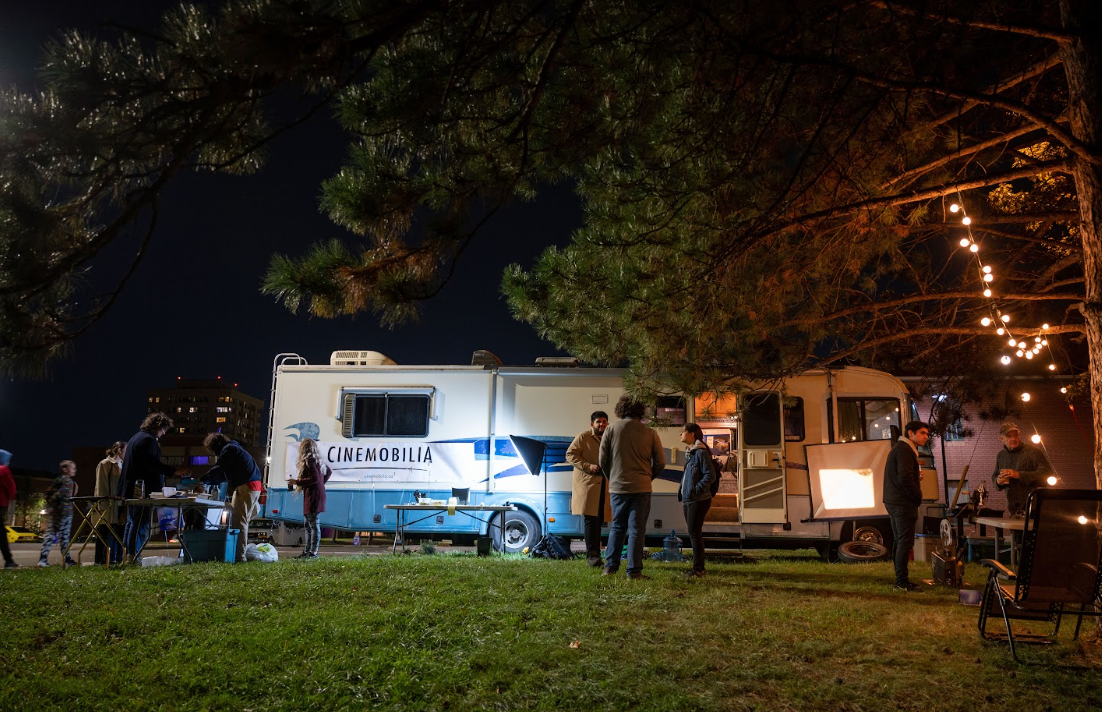 A photograph of a group of young people standing in front of an RV at night with lots of film lights and tables around them.