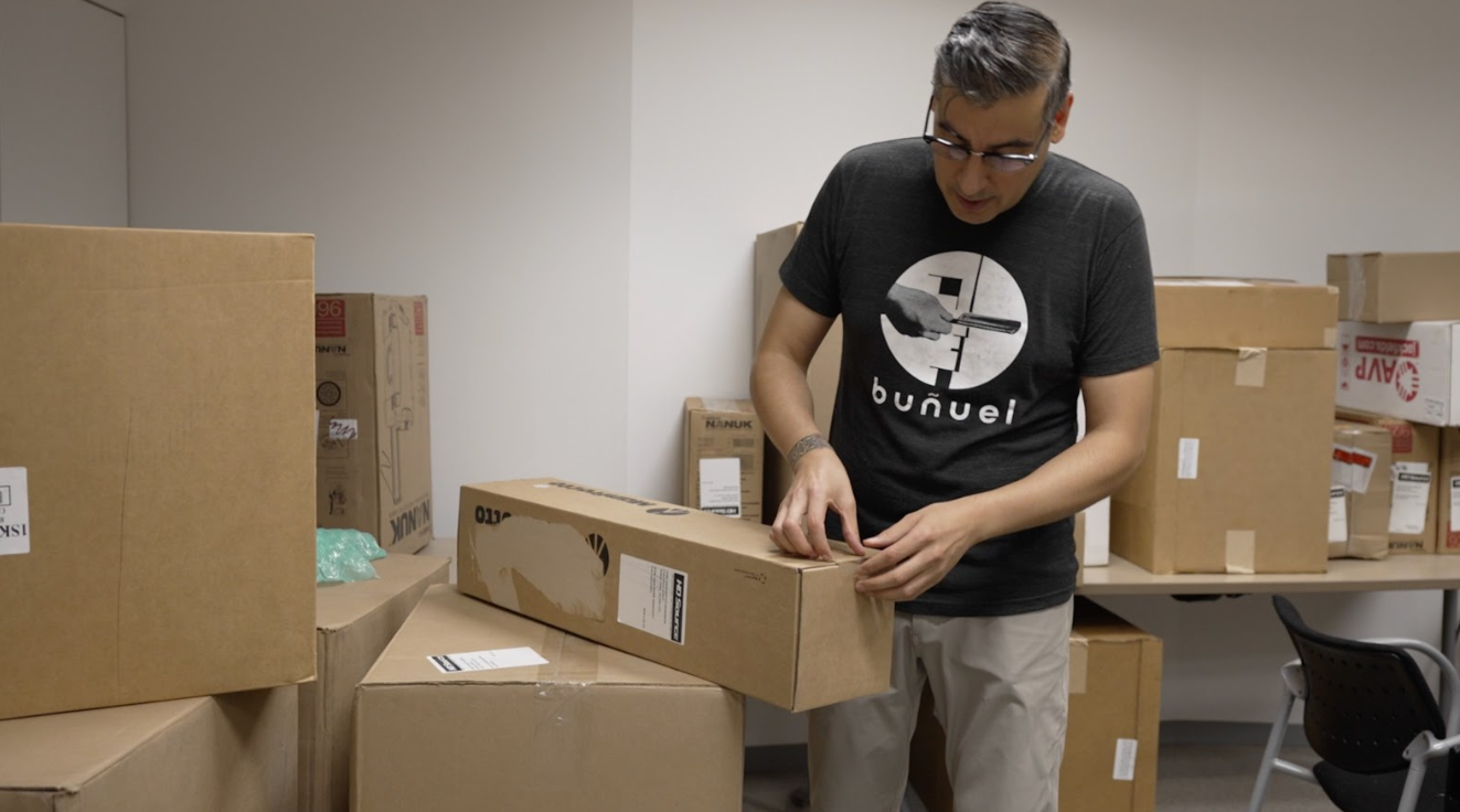A photograph of a man with black hair, a black printed t-shirt, and gray pants who is opening up a bunch of cardboard boxes.