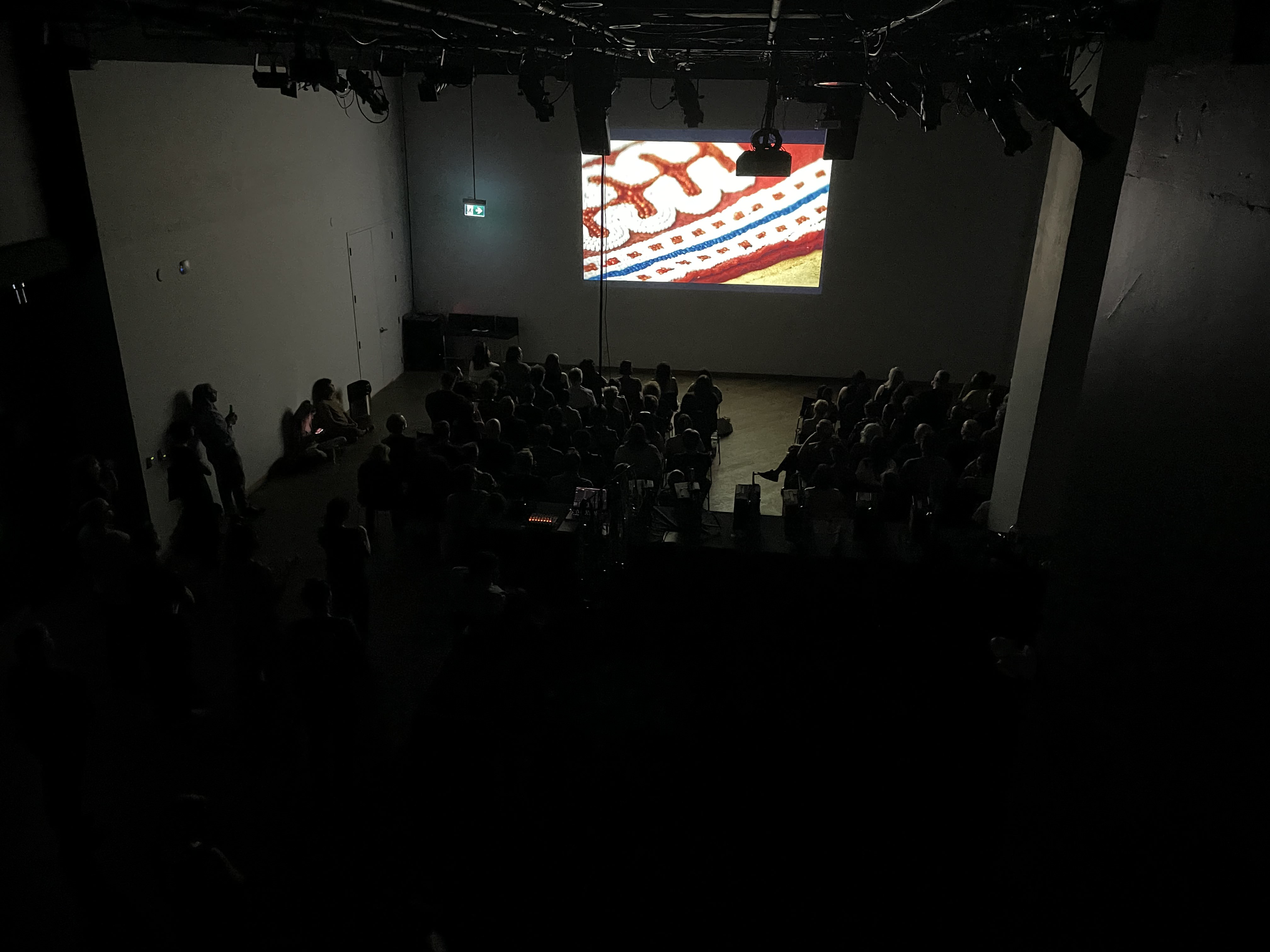 A photograph taken from a high angle of a dark venue space with lots of people sitting in a grid of chairs watching a digital projection.
