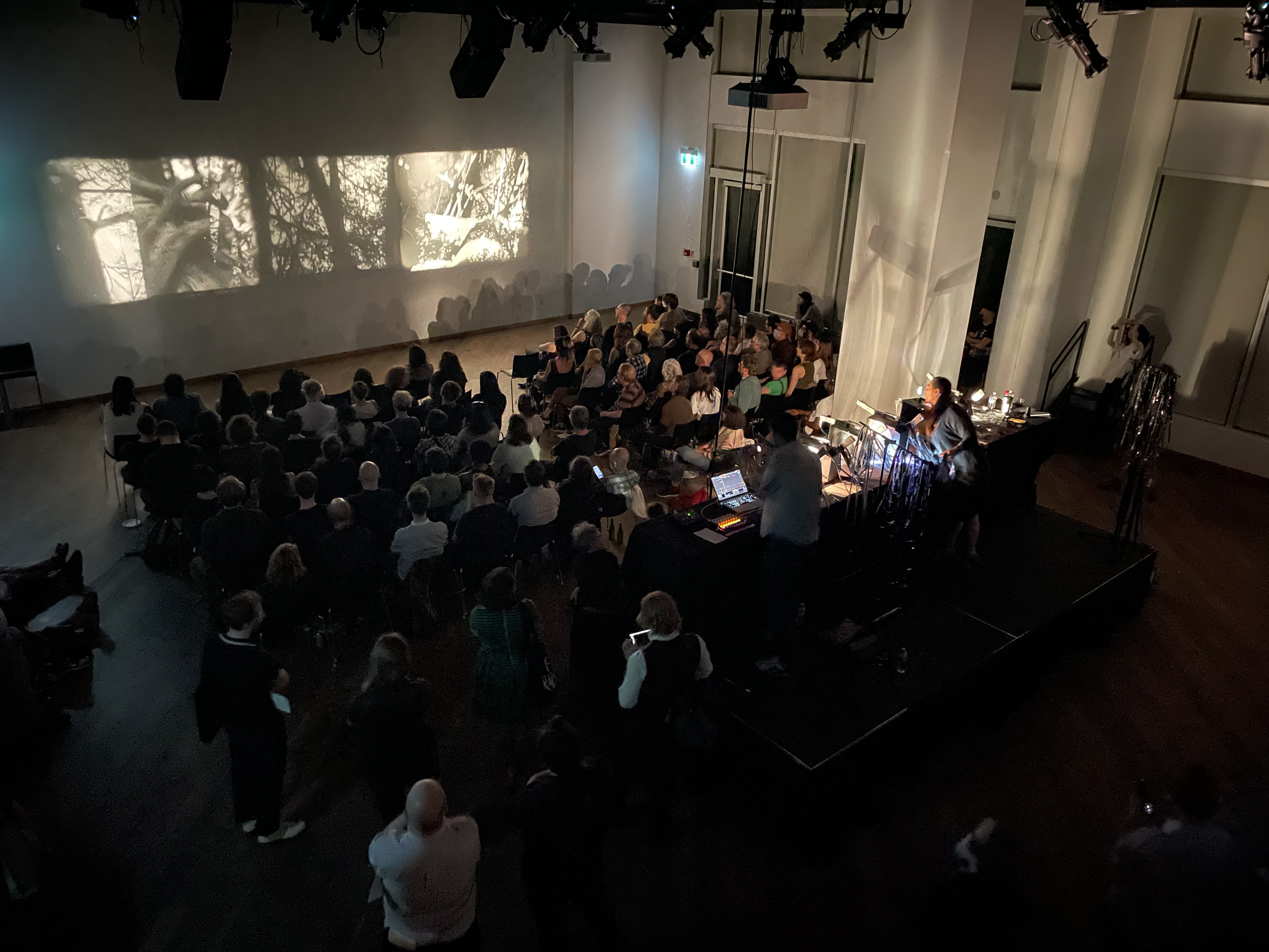 A photograph taken from a high angle of a dark venue space with lots of people sitting in a grid of chairs watching an analog film projection.