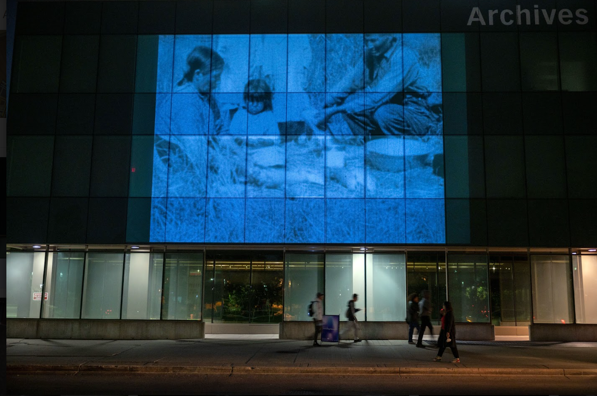 A photograph of a large video of some people sitting under a tree being projected onto the side of a building at night.