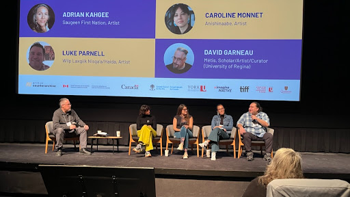 A photo of three women and two men sitting on lounge chairs on a theatre stage. There is a large digital slide projection behind them and they are all holding microphones.