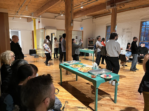 A photograph of a group of people standing and sitting in a room with high wooden ceilings and polishing wooden floors. There are a couple of teal tables with food arranged around the space and most fo the people seem to be looking toward something happening outside of the frame on the right side of the photo.