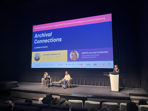 A theatre stage with two women sitting in chairs and another woman standing at a podium. There is a large digital slide projection behind them that displays the text "Archival Connections" on it.