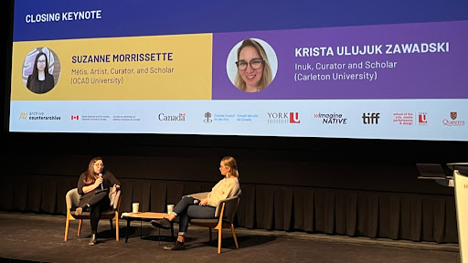 A theatre stage with two women sitting in chairs. There is a large digital slide projection behind them that displays the text "Archival Connections" on it.