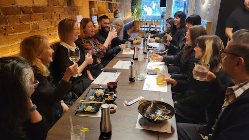 A photograph of a large group of people eating in a sushi restaurant and who are all looking toward a woman in the centre raising their glass in a celebratory cheer.