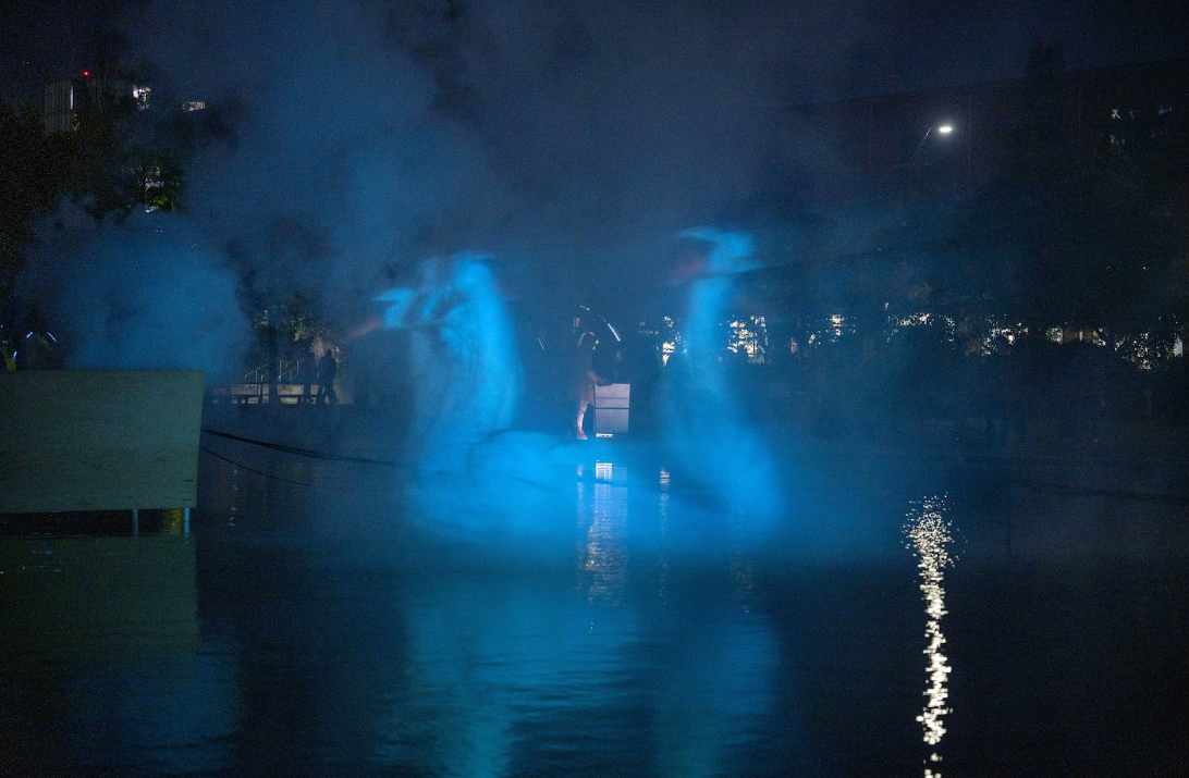 A photograph of a large artificial pond with a video of a group white swans being projected onto a cloud of fog above the water at night.