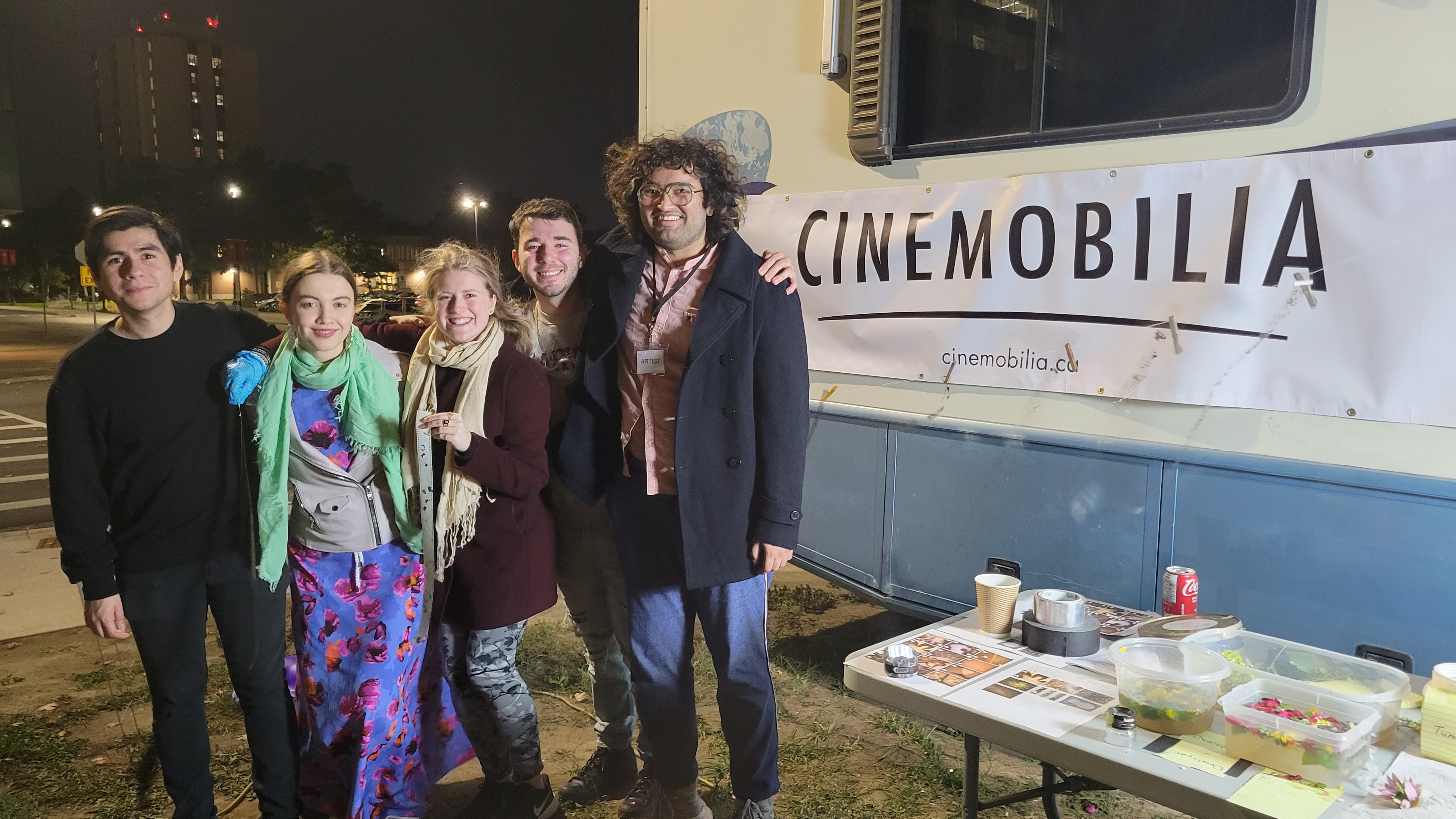 A photograph of a group of young people standing in front of an RV at night with lots of film lights and tables around them.