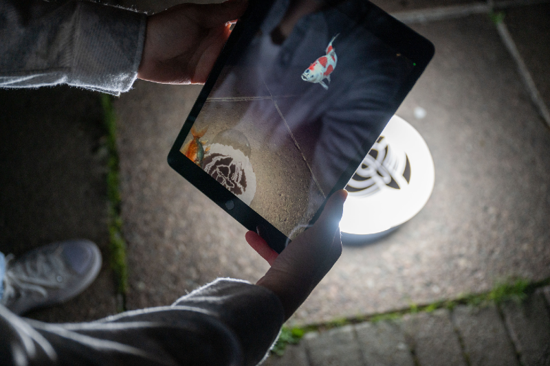 A colour photograph taken at night of a person's hands holding a digital tablet. The tablet is in camera mode showing an AR depiction of some fishing swiming in the air above the ground where the tablet is being aimed at.