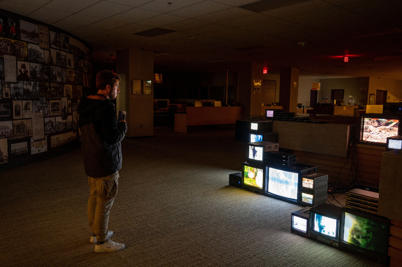 A colour photograph of a dimly lit institutional space containing a media art installation consisting of a number old televisions with footage of chickens being shown. There is also a young man looking at the installation who is holding a cup of coffee.