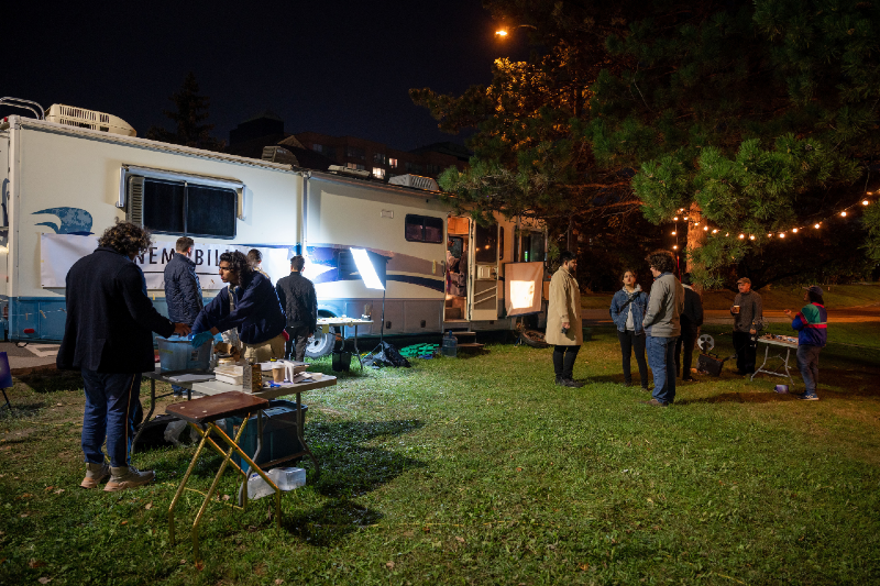 A colour photograph taken at night of a large white trailer van with a hanging bulb lights and tables set up around it. There are many people meandering around the tables chatting with one another.