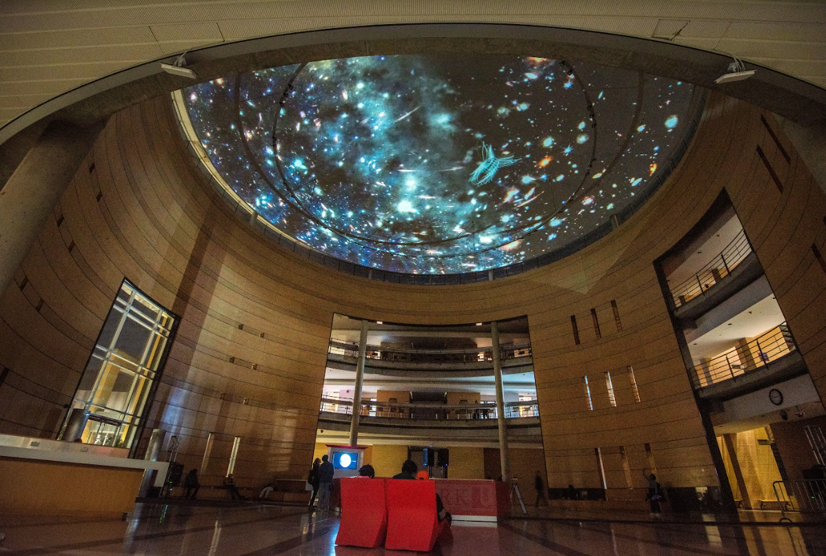 A photograph of a lobby space where a large video projection of the night sky is being projected onto a domed ceiling.
