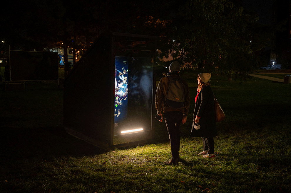 A photograph of two people standing in front of a large booth with a video monitor mounted vertically inside of it. The booth has light projecting from it and is installed in a lightly forested area at night.