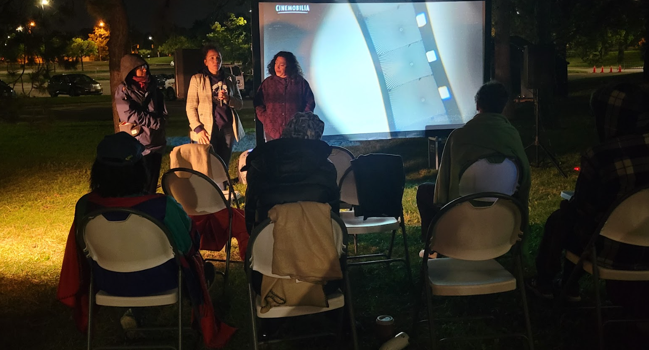A photograph of an outdoor movie screening at night in a slightly forested area. There are three people speaking in front of the screen and a bunch of people sitting in folder chairs watching them.