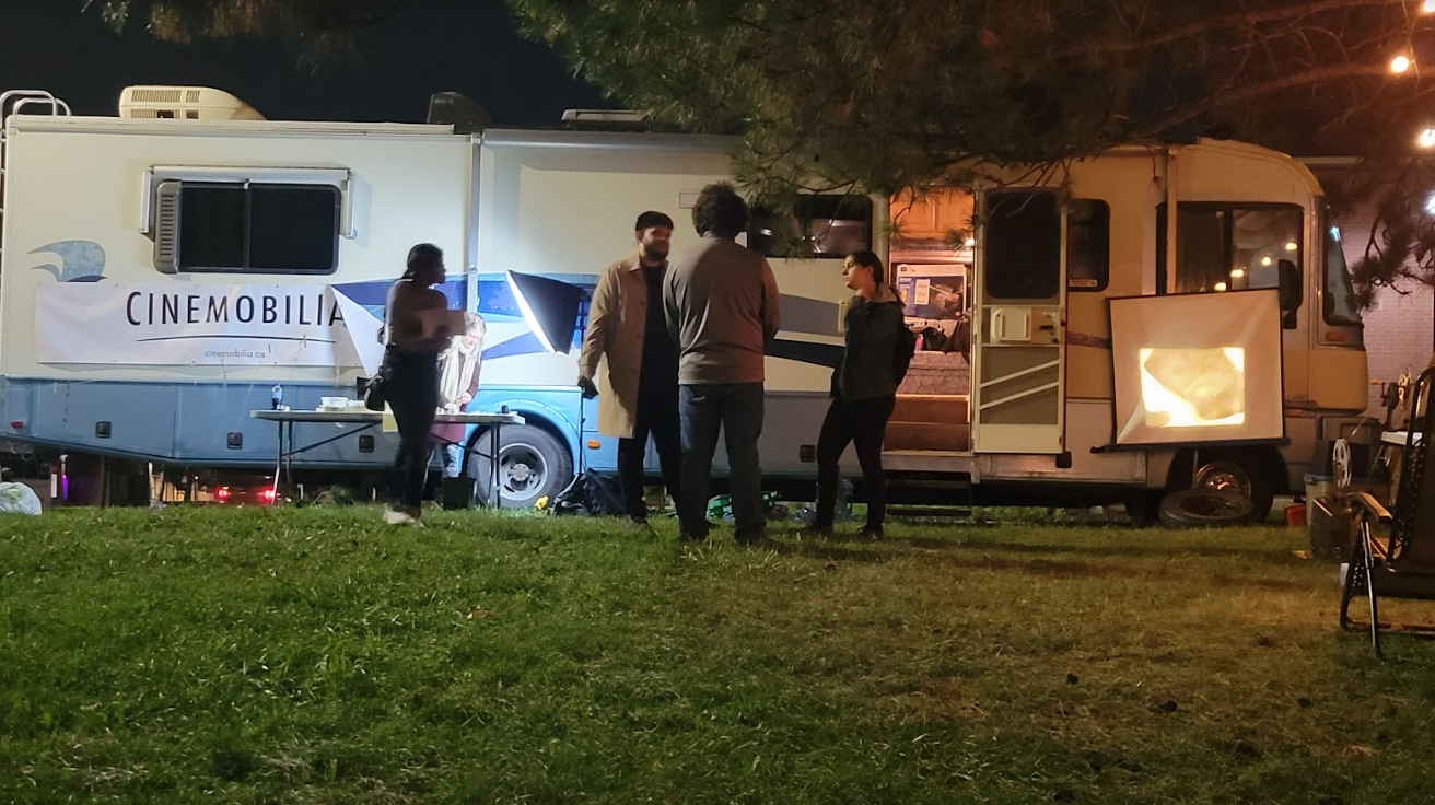A photograph of a group of people standing outside of an RV at night with film lighting and tables scattered around.