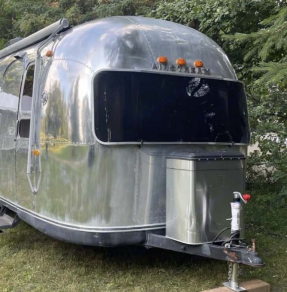 A photograph of a silver metallic camper parked in front of some trees and resting on some grass.