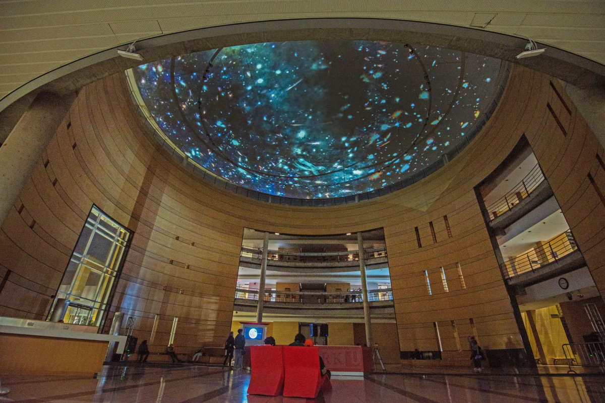 A colour photograph of a an institutional university lobby space where a large video of a starry night sky is being digitall projected on a semi-spherical domed ceiling.