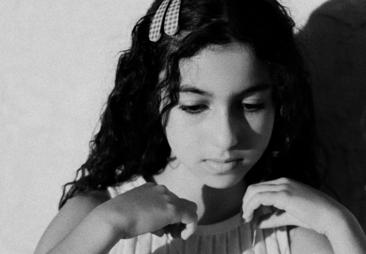 A black and white film still of a young woman with long black hair holding her hands up to her shoulders.