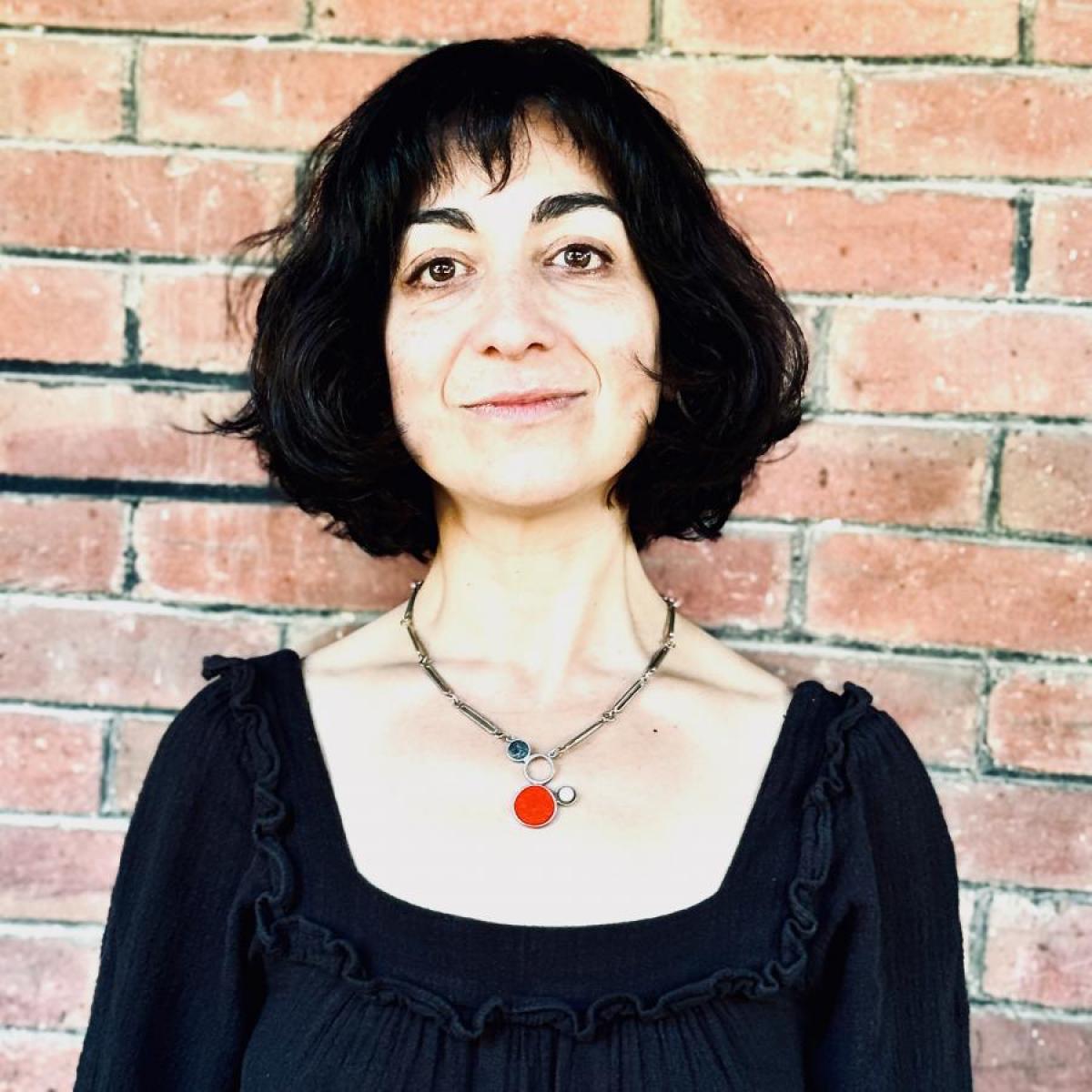 Medium shot of Erika Biddle, new A/CA KMO, with short dark hair, in a black dress, with a red necklace, against a brick wall