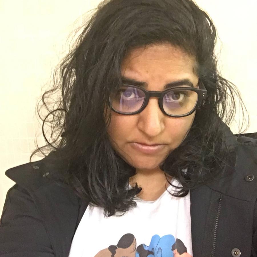 The image depicts Pamila Matharu. She is wearing a white shirt with figures on it, and a black jacket. Matharu has brown skin, wears glasses, and has medium long hair.