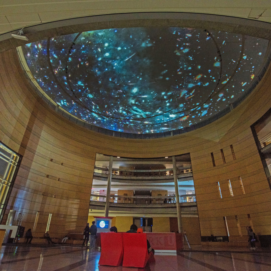 A colour photograph of a an institutional university lobby space where a large video of a starry night sky is being digitall projected on a semi-spherical domed ceiling.