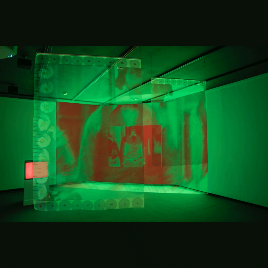 A colour photograph of a dimly lit gallery space with large sheets of semi-transluscent fabric hanging from the ceiling. The fabric have slight abstracted red digital prints on them and the gallery is lit using lime green lighting.