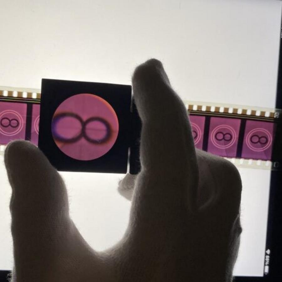 A promotional image for the Eye International Conference 2023 that depicts a persons hand holding a small sqaure magnifying glass over a piece of old analog film on top of a light table. The film strip is showing many frames of a the number 8 shown in black against a purple background.