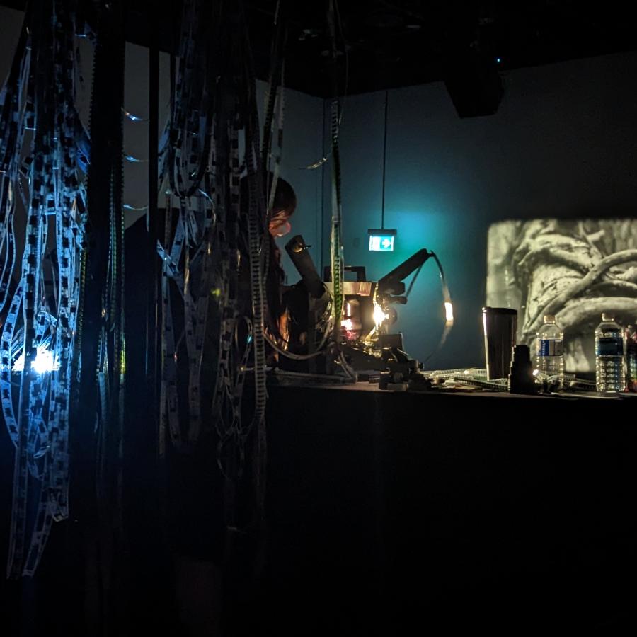 A photograph taken of a woman operating several analog film projectors in a dark space. There is a black and white projection in front of her and long strands of film reel hanging from behind her.