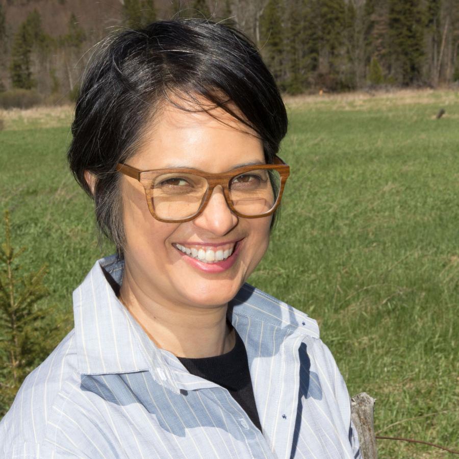 A portrait photograph of smiling woman with dark hair, brown glasses, and a white collar shirt who is standing out in a grassy field on a sunny day.