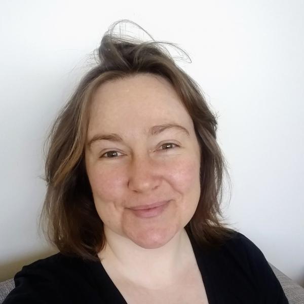 A photograph of a woman with light brown hair and a black shirt sitting against a white wall and smiling.