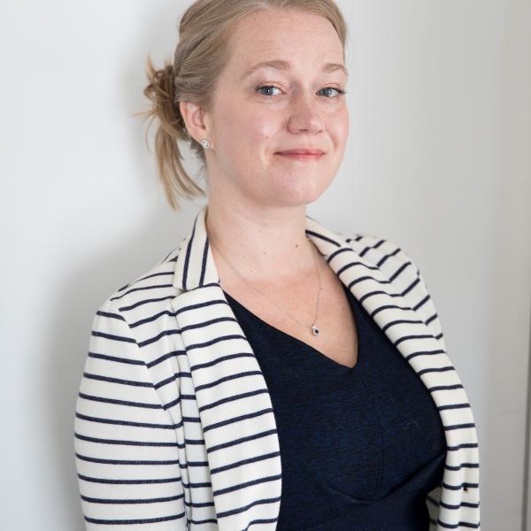 A photo of a woman with a blond ponytail leaning against a white wall. She is wearing a black and white striped cardigan and a black shirt.