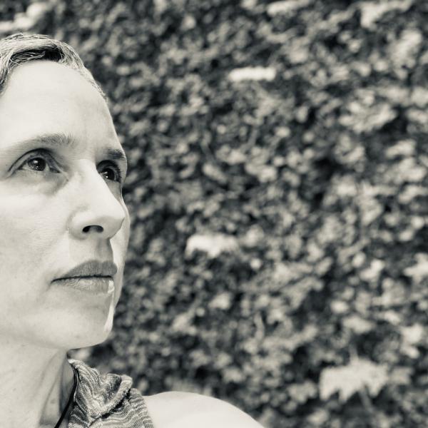 A black and white portrait photo of a woman looking slightly upward and standing in front of a large wall with vines and leaves on it.