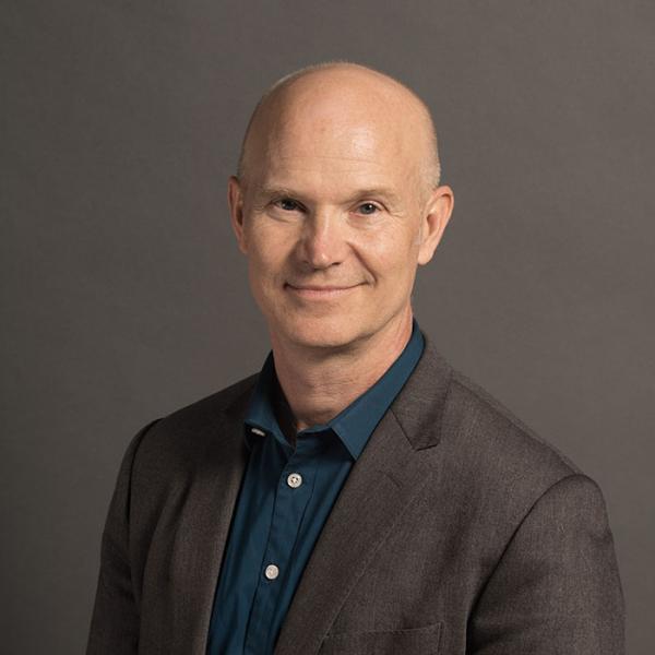 A photograph of a man in a dark grey suit and blue collared shirt standing in front a dark grey background.