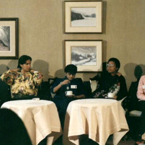 A film still of a group of women sitting around small circular tables in front of a wall with four large framed pieces of art hung on it.