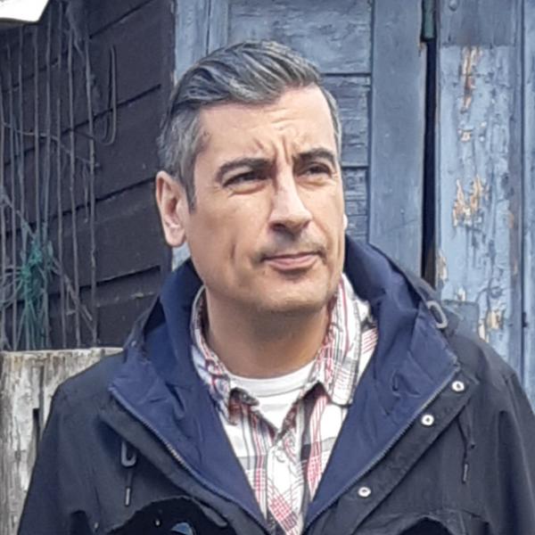 A photograph of a man with slicked back hair, wearing a navy coat and a plaid collared shirt standing in front of a faded blue wooden wall.