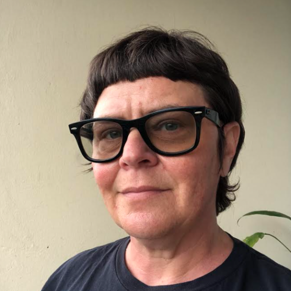 A photograph a person with medium length dark brown hair and tinted glasses standing in front of a blank beige wall and a plant.