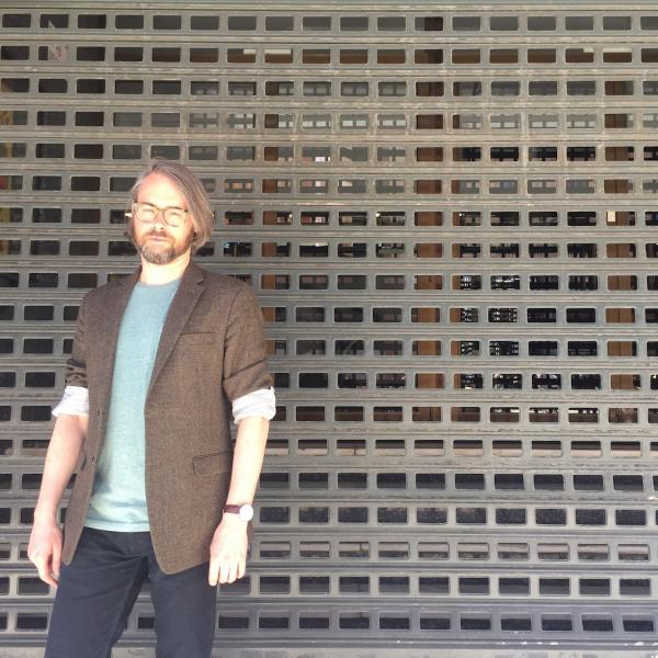 A photograph of a man with medium length hair, a beard, glasses, and a blazer who is standing in front of a outdoor gridded wall during a bright day.