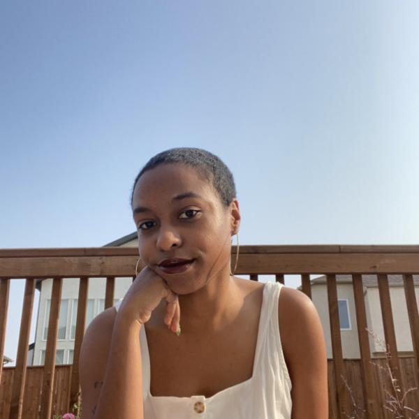 A photograph of a young woman sitting in front a brown wooden railing with the roofs of some houses and a clear blue sky behind them.