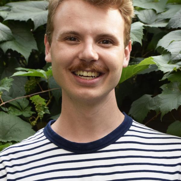 A photo of a smiling young man with a striped shirt, a moustache, and short light brown hair standing in front of a bush.