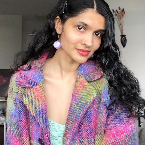 A photograph of a woman with a pink and purple coat and large circular white earrings who is standing in an interior space with white walls.